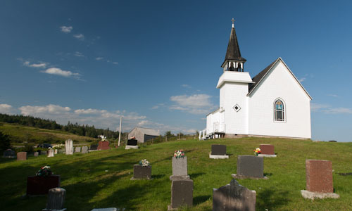 A photo of the rear of a small white church surrounded on two sides by headstones