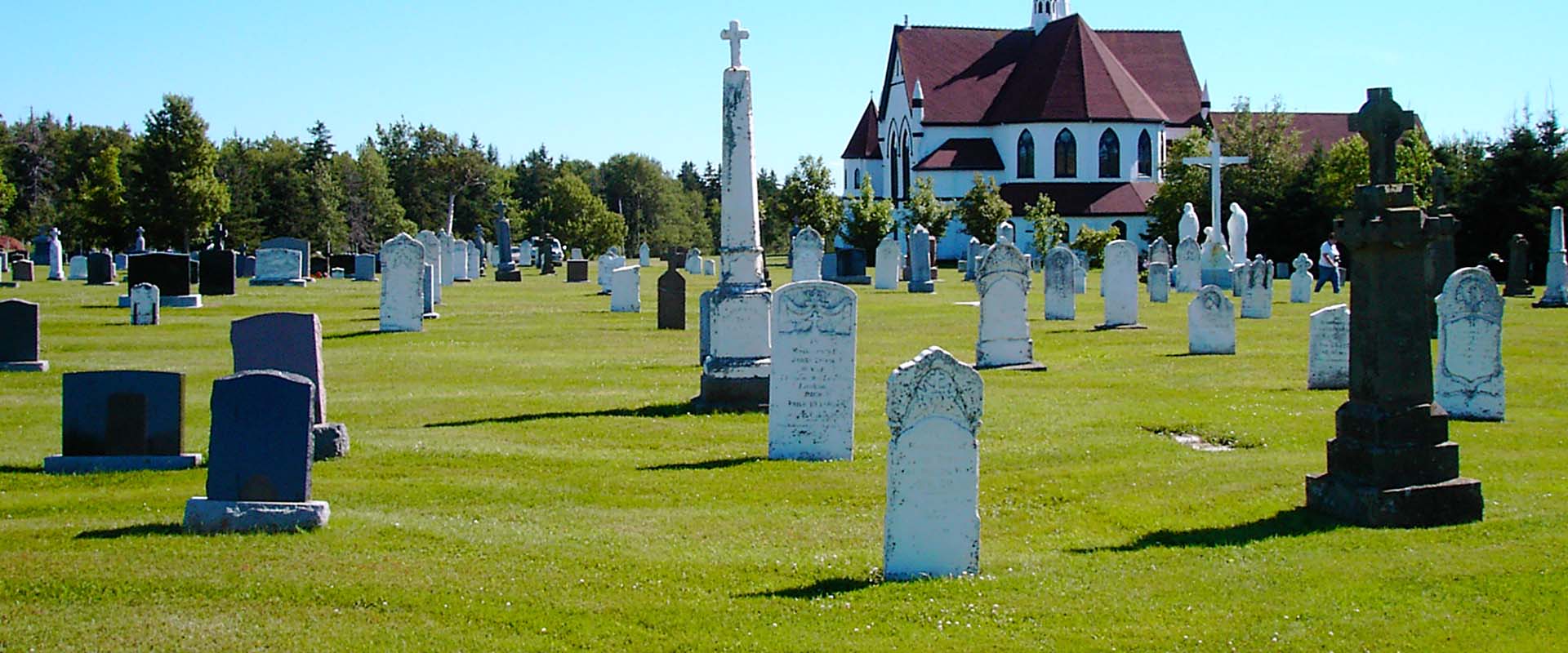 A view of headstones located to the side and rear of a wooden country church. The image displays the cemetery and church of St. Mary's Roman catholic parish in Indian River, Prince Edward Island.